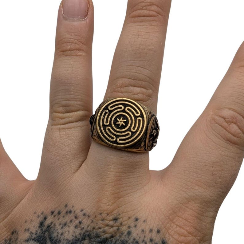 May include: A gold-toned metal ring featuring a circular face with a raised labyrinth pattern and a central star-like symbol. The band has a textured, dark finish on the sides, providing a contrast to the polished gold-colored surface of the ring.