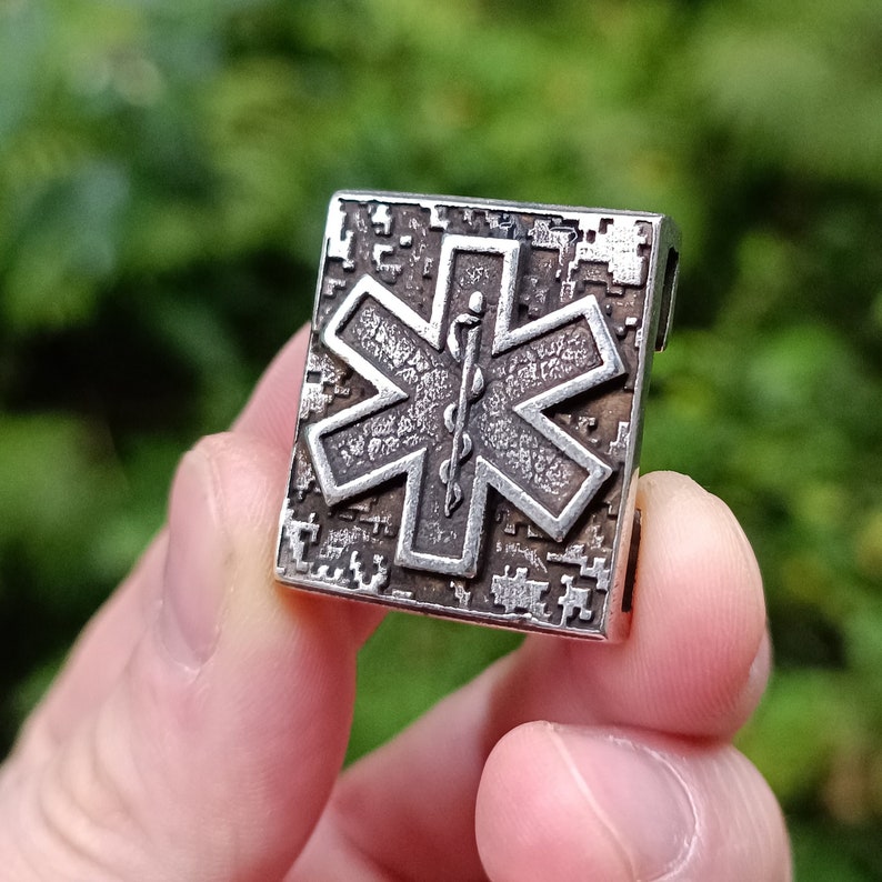 Pode incluir: Um amuleto de metal prateado com um design detalhado de estrela da vida. A estrela da vida &eacute; um s&iacute;mbolo usado pelos servi&ccedil;os m&eacute;dicos de emerg&ecirc;ncia.