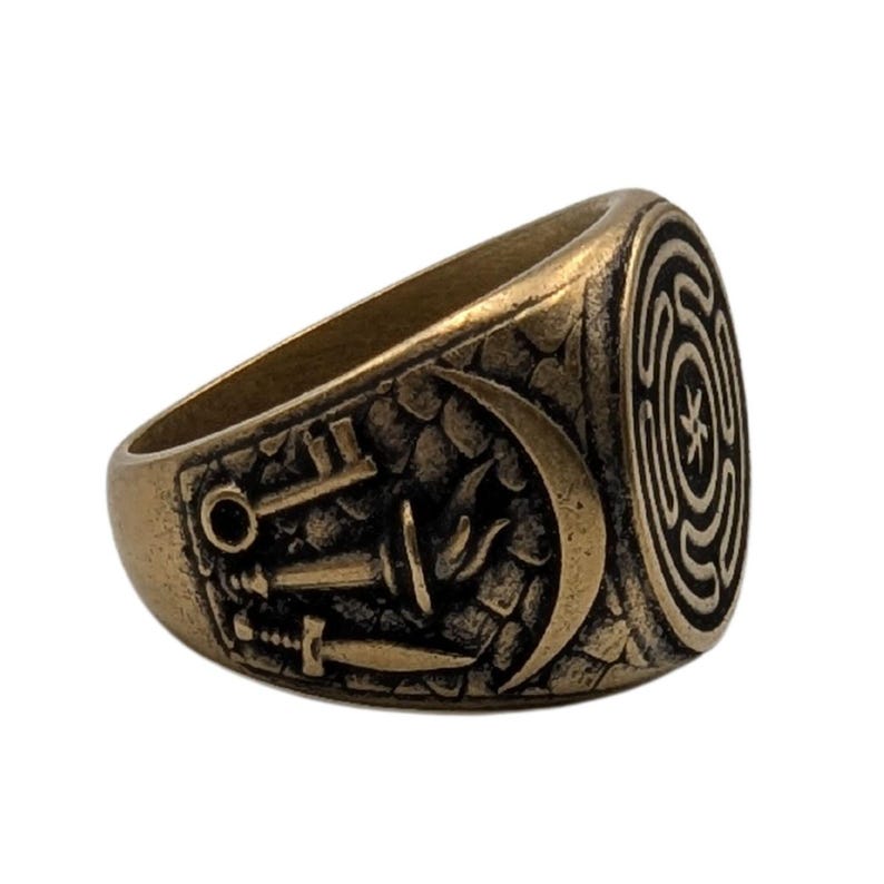 May include: A brass-toned metal ring featuring an oval face with a central star emblem surrounded by concentric lines. The side of the band displays a relief design of a key, a torch, and a sword set against a textured background of stone-like blocks.