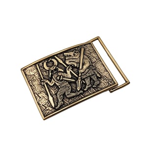 May include: A rectangular brass-toned belt buckle featuring a raised relief design of a horned figure holding a spear and a wolf-like creature standing opposite, both depicted in a textured, antique-style finish with a metal loop on the side.
