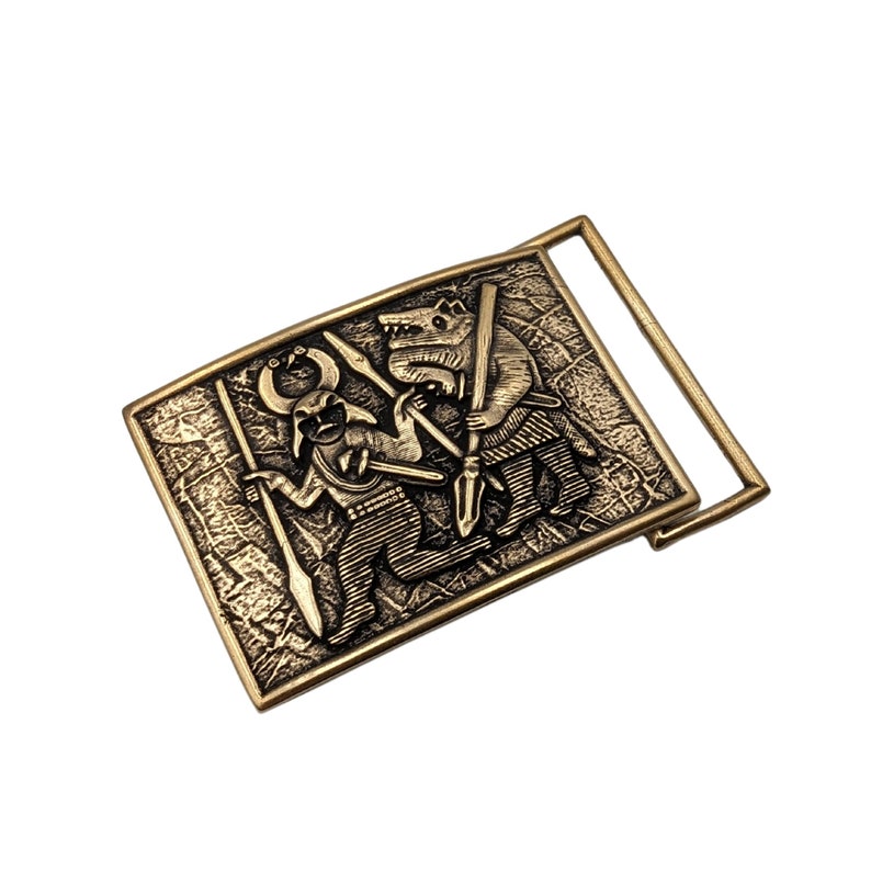 May include: A rectangular brass-toned belt buckle featuring a raised relief design of a horned figure holding a spear and a wolf-like creature standing opposite, both depicted in a textured, antique-style finish with a metal loop on the side.