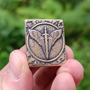 Könnte beinhalten: Ein quadratischer Metalls Anhänger mit einer strukturierten Oberfläche. Der Anhänger zeigt ein kreisförmiges Design mit einem Schwert und Flügeln. Das Schwert ist silberfarben und die Flügel sind golden.