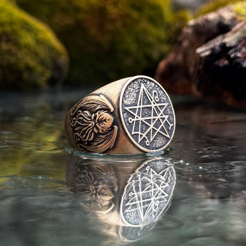 Może przedstawiać: Metalowy pierścień o wykończeniu z antycznego brązu z okrągłą tarczą wygrawerowaną w złożony siedmioramienny symbol gwiazdy. Boki pierścienia przedstawiają szczeg&oacute;łowy relief uskrzydlonego stworzenia z mackami o wyraźnej teksturze powierzchni.