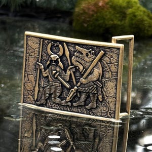 May include: A rectangular brass-toned belt buckle featuring a relief design of a horned figure holding a spear alongside a wolf-like creature holding a spear. The background has a textured, weathered appearance with a metallic finish.