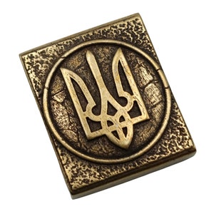 May include: A gold-colored metal square with a raised, circular design featuring the Ukrainian trident symbol.