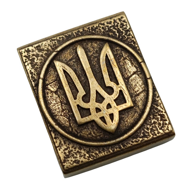 May include: A gold-colored metal square with a raised, circular design featuring the Ukrainian trident symbol.