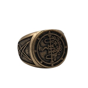 May include: A gold-toned ring with a black sigil design. The sigil is a circular design with intricate lines and symbols.