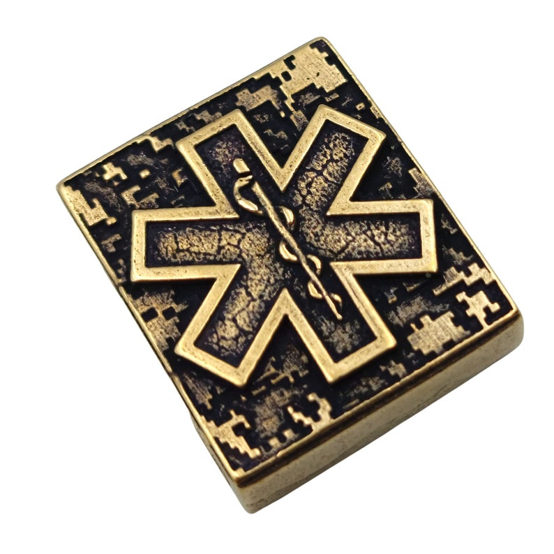 Pode incluir: Um amuleto de metal dourado com um padr&atilde;o de camuflagem preto e dourado. O amuleto apresenta um s&iacute;mbolo m&eacute;dico estilizado em relevo com uma cobra enrolada em torno de um cajado.