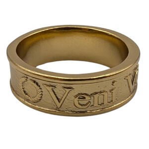 May include: A gold-toned metal band ring featuring a textured surface with the embossed text Veni and a laurel wreath symbol. The ring has a smooth interior and a consistent width throughout, showcasing a metallic finish with visible engraved details.