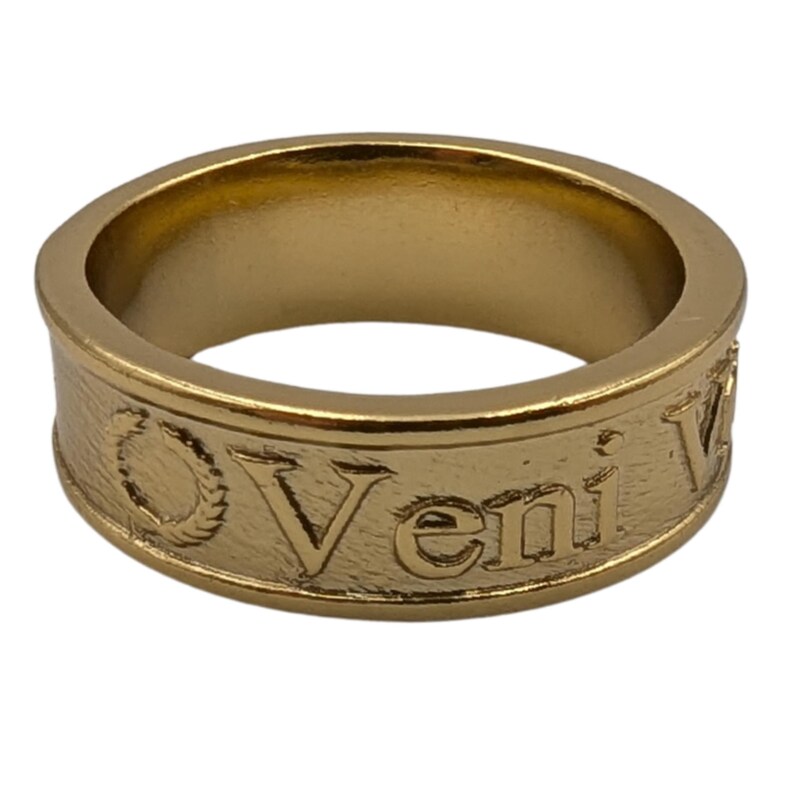May include: A gold-toned metal band ring featuring a textured surface with the embossed text Veni and a laurel wreath symbol. The ring has a smooth interior and a consistent width throughout, showcasing a metallic finish with visible engraved details.