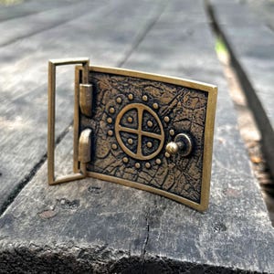 May include: A rectangular brass belt buckle with a dark, weathered finish. The surface features a central circular motif divided into four quadrants, surrounded by a ring of small raised dots. A metal loop and a small stud are attached to the side.