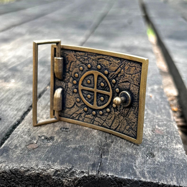 May include: A rectangular brass belt buckle with a dark, weathered finish. The surface features a central circular motif divided into four quadrants, surrounded by a ring of small raised dots. A metal loop and a small stud are attached to the side.