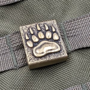 Könnte beinhalten: Ein Messing-Bärenpfoten-Anhänger mit strukturierter Oberfläche. Der Anhänger ist an einem braunen Stoffband befestigt.