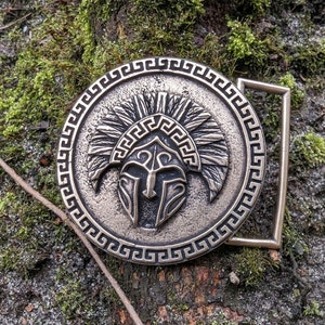 May include: A circular bronze-toned belt buckle featuring a raised relief of a Spartan helmet with a plume. The outer edge of the buckle is decorated with a repeating Greek key pattern. A rectangular metal loop is attached to the right side.