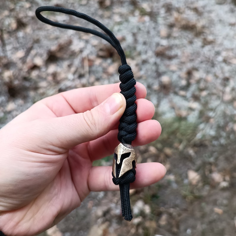 May include: A black paracord lanyard with a bronze Spartan helmet bead. The lanyard is tied in a knot and is held in a hand.
