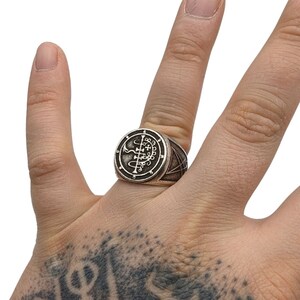 May include: A silver ring with a circular design featuring a sigil and a pentagram. The ring is worn on a finger.