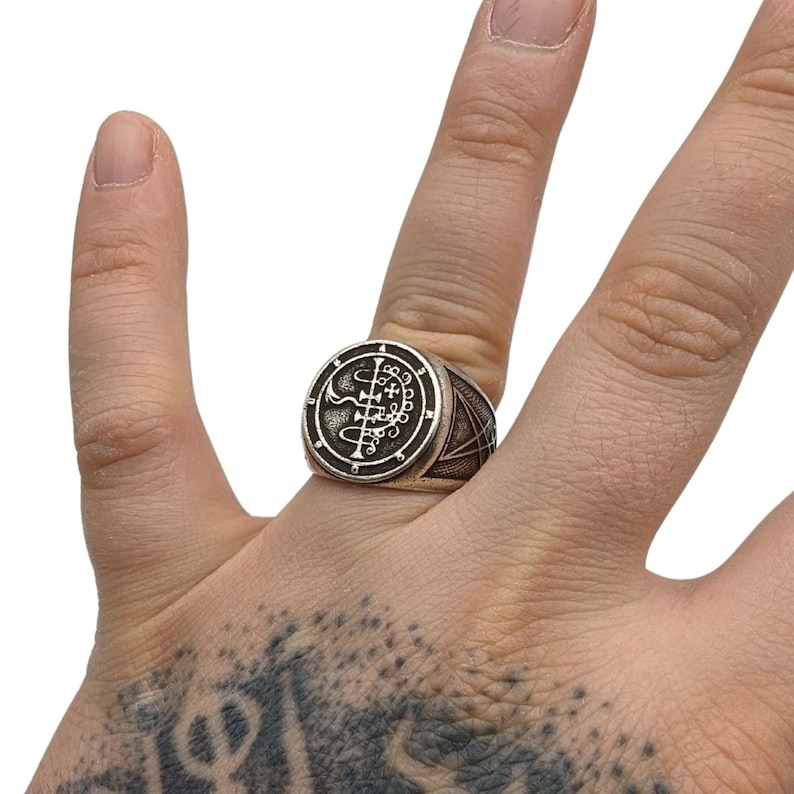 May include: A silver ring with a circular design featuring a sigil and a pentagram. The ring is worn on a finger.