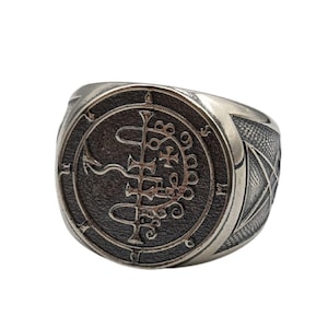 May include: A silver ring with a black sigil design. The sigil is a circle with a cross and other symbols inside. The ring is a statement piece for those who appreciate unique jewelry.