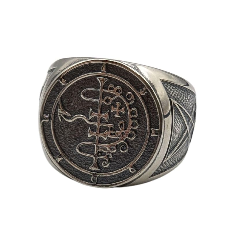 May include: A silver ring with a black sigil design. The sigil is a circle with a cross and other symbols inside. The ring is a statement piece for those who appreciate unique jewelry.