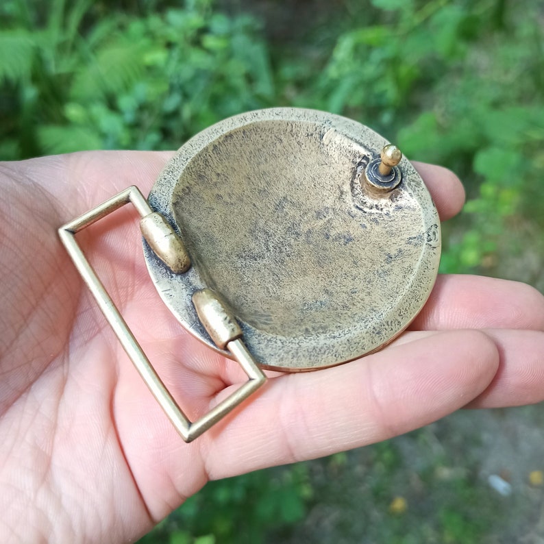 May include: A circular brass belt buckle with a textured, weathered finish. The buckle features a rectangular frame attached to the side and a small, upright metal post on the back surface for securing the belt strap in place.