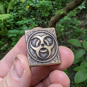 May include: A brass square-shaped pendant with a textured surface and a raised biohazard symbol in the center.