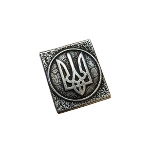 May include: A silver metal pin with a textured surface featuring the Ukrainian coat of arms, a trident, in a circle.