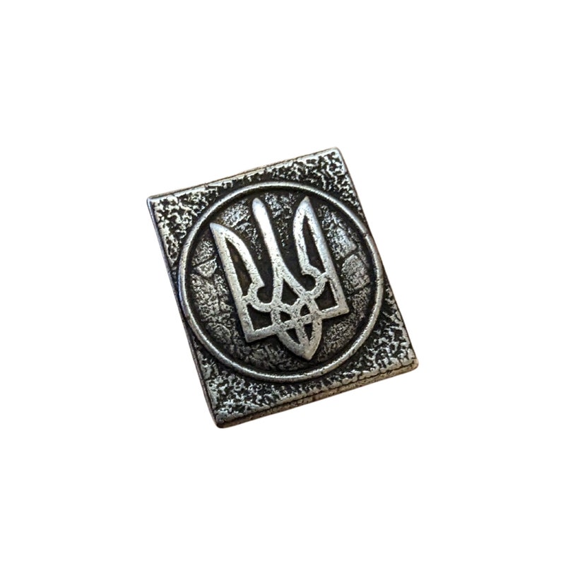 May include: A silver metal pin with a textured surface featuring the Ukrainian coat of arms, a trident, in a circle.