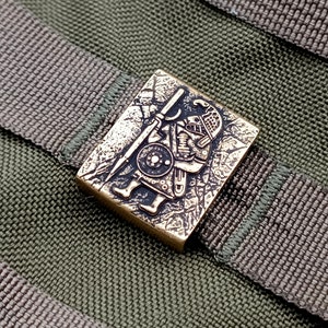 Könnte beinhalten: Eine bronzene Metallstifte mit einem detaillierten Design eines Kriegers, der eine Lanze und einen Schild hält. Der Stift befindet sich auf einem grünen Stoffhintergrund.