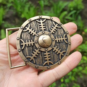 May include: A bronze belt buckle with a circular design featuring a stylised sun with eight rays. The buckle has a textured, wood-like finish and a raised centre.