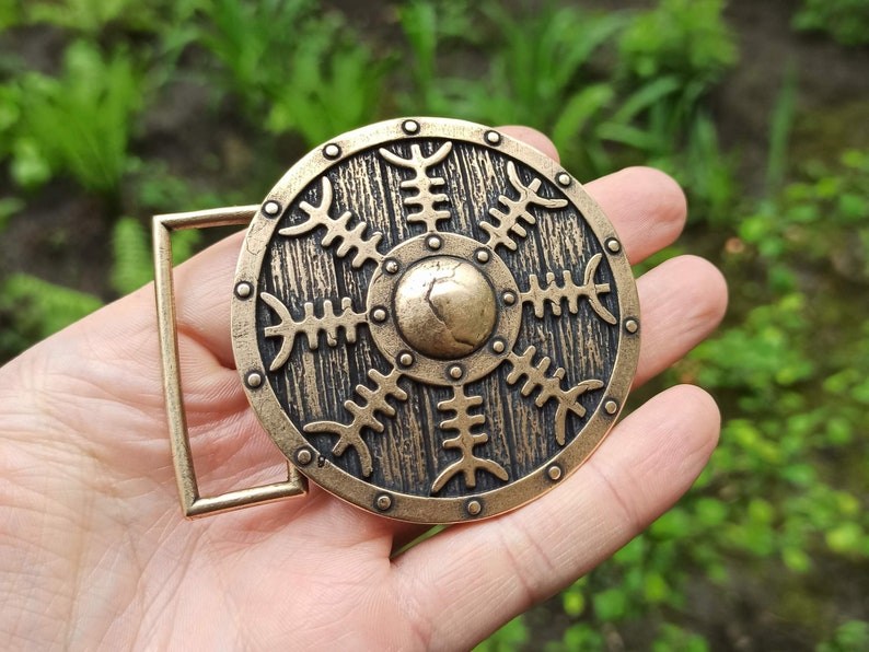 May include: A bronze belt buckle with a circular design featuring a stylised sun with eight rays. The buckle has a textured, wood-like finish and a raised centre.