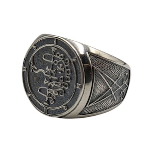 May include: A silver ring with a round, engraved design featuring a sigil and a circle of numbers. The band of the ring is also engraved with a geometric pattern.