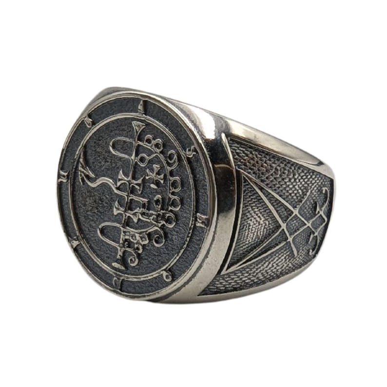 May include: A silver ring with a round, engraved design featuring a sigil and a circle of numbers. The band of the ring is also engraved with a geometric pattern.