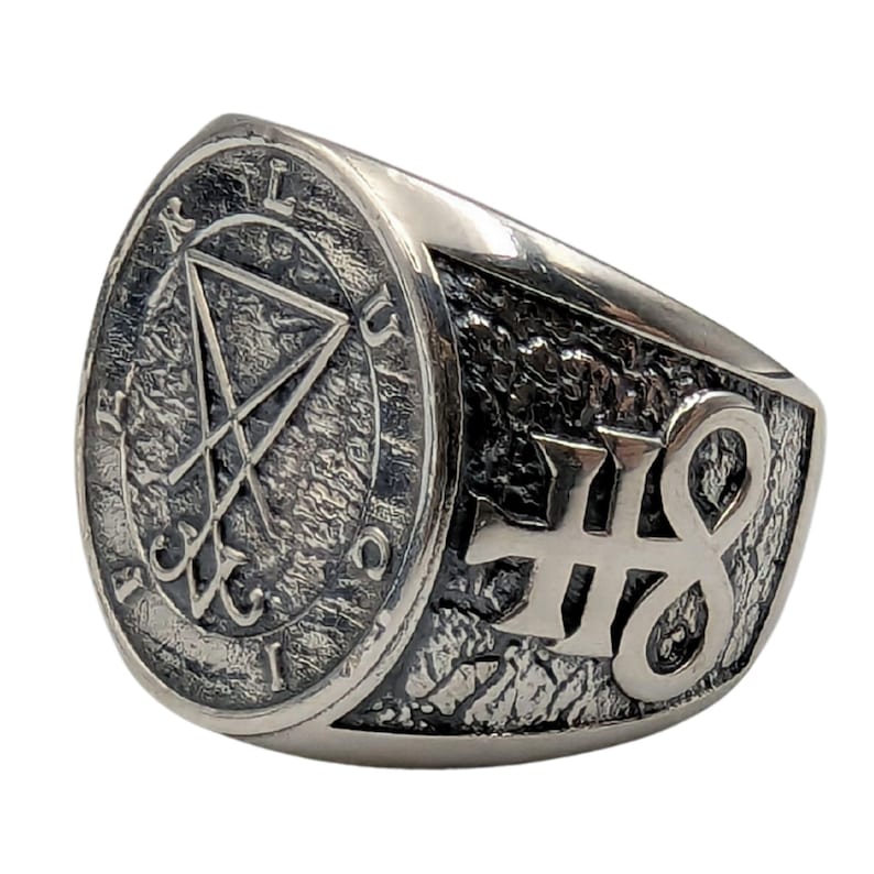 May include: A silver-toned metal ring featuring a textured, oxidized finish. The circular face displays a sigil of Lucifer enclosed in a circle with the letters L, U, C, I, F, E, R inscribed around the border. The side features a raised symbol.