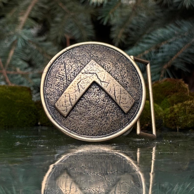 May include: A circular bronze-toned belt buckle featuring a raised, inverted V-shaped emblem in the centre. The surface has a distressed, weathered texture with dark patina accents throughout the metal, and a rectangular loop attached to the side.