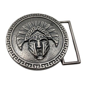 May include: A circular silver-toned metal belt buckle featuring a raised relief of a crested helmet in the centre. The outer edge of the circular design is decorated with a repeating geometric Greek key pattern, and a rectangular loop is attached.