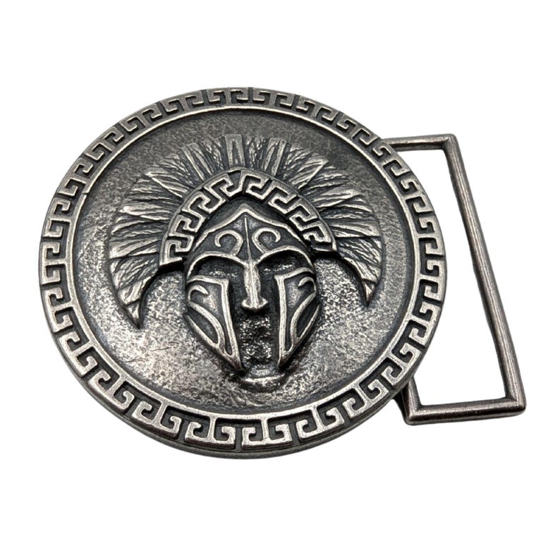 May include: A circular silver-toned metal belt buckle featuring a raised relief of a crested helmet in the centre. The outer edge of the circular design is decorated with a repeating geometric Greek key pattern, and a rectangular loop is attached.