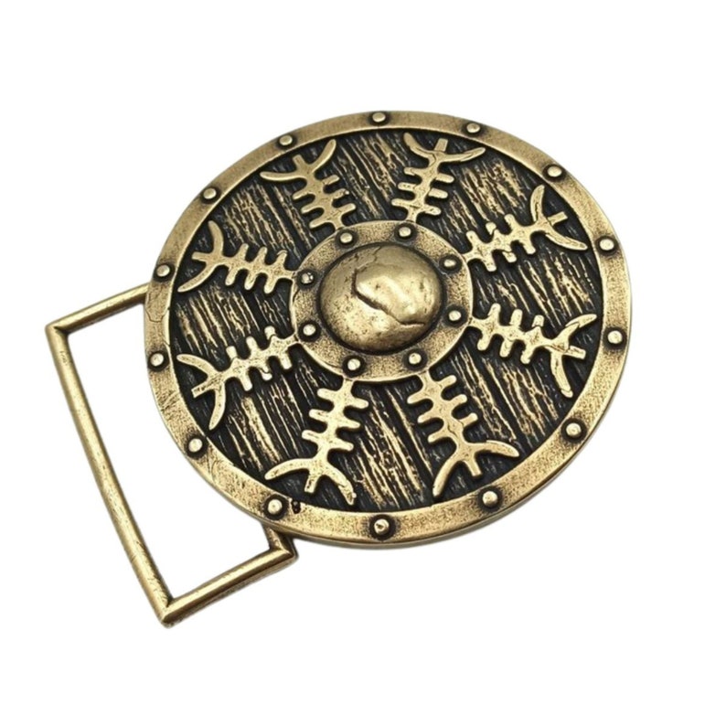 May include: A gold-toned belt buckle featuring a round shield design. The shield has a black and gold pattern and a raised centre. The buckle is attached to a metal loop.