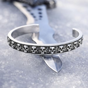 May include: Silver cuff bracelet featuring a repeating skull design. The bracelet is open at the ends and has a detailed, textured surface. The skulls are evenly spaced around the band, creating a gothic or edgy aesthetic.