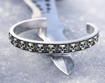 May include: Silver cuff bracelet featuring a repeating skull design. The bracelet is open at the ends and has a detailed, textured surface. The skulls are evenly spaced around the band, creating a gothic or edgy aesthetic.