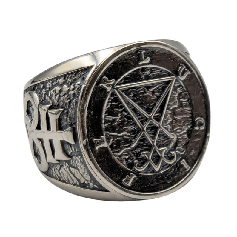 May include: A silver-toned metal ring featuring a circular face with a textured dark background. The center displays a geometric sigil, surrounded by the letters L, U, C, I, F, E, R. The sides of the band are decorated with an embossed cross symbol.