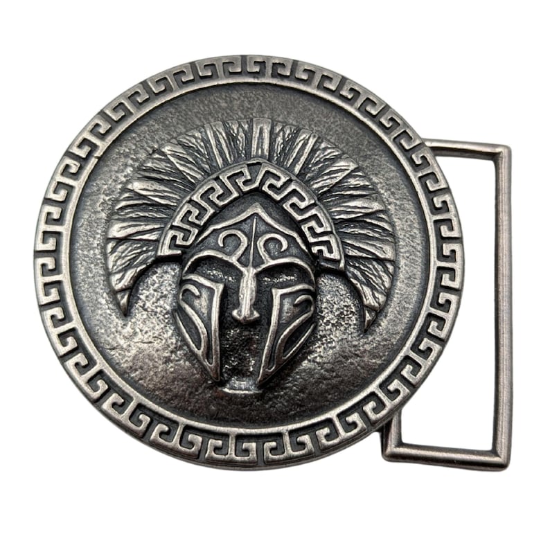 May include: A circular silver-toned metal belt buckle featuring a raised relief of a crested helmet in the centre. The outer edge of the buckle is decorated with a repeating geometric Greek key pattern, and a rectangular loop is attached to the side.