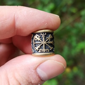 May include: A black and gold metal bead with a Vegvisir symbol, a Norse directional symbol, engraved on it.
