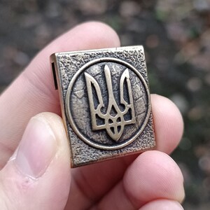 May include: A gold-colored metal charm with a detailed depiction of the Ukrainian trident, a three-pronged symbol, in a circular design.