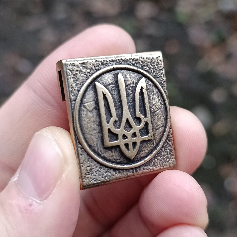 May include: A gold-colored metal charm with a detailed depiction of the Ukrainian trident, a three-pronged symbol, in a circular design.