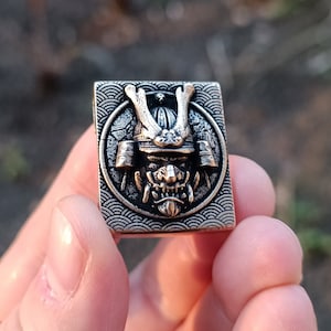 May include: A silver and black metal pendant featuring a detailed, three-dimensional samurai warrior's face with a fierce expression. The pendant is surrounded by a decorative wave pattern.