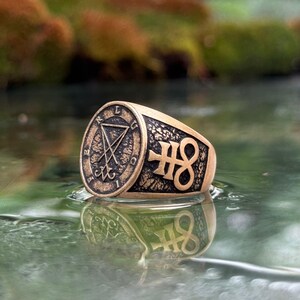 May include: A bronze-toned metal ring featuring a circular face with the Sigil of Lucifer and the letters L, U, C, I, F, E, R engraved around the border. The side of the ring displays a raised Leviathan Cross symbol against a textured background.