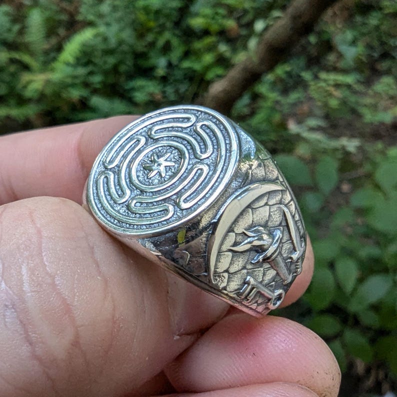 May include: A silver-toned ring featuring a circular labyrinth design with a central star motif on the face. The side of the band displays an engraved crescent moon, a torch, and a key set against a textured, scale-like background pattern.
