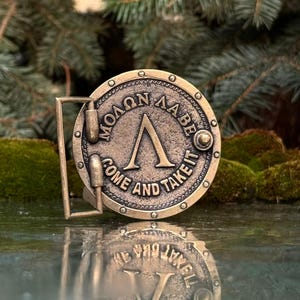 May include: A circular brass-toned metal belt buckle featuring a raised Greek Lambda symbol in the centre. The text "&Mu;&Omicron;&Lambda;&Omega;&Nu; &Lambda;&Alpha;&Beta;&Epsilon;" and "COME AND TAKE IT" is embossed around the perimeter, which is accented with small circular rivets.
