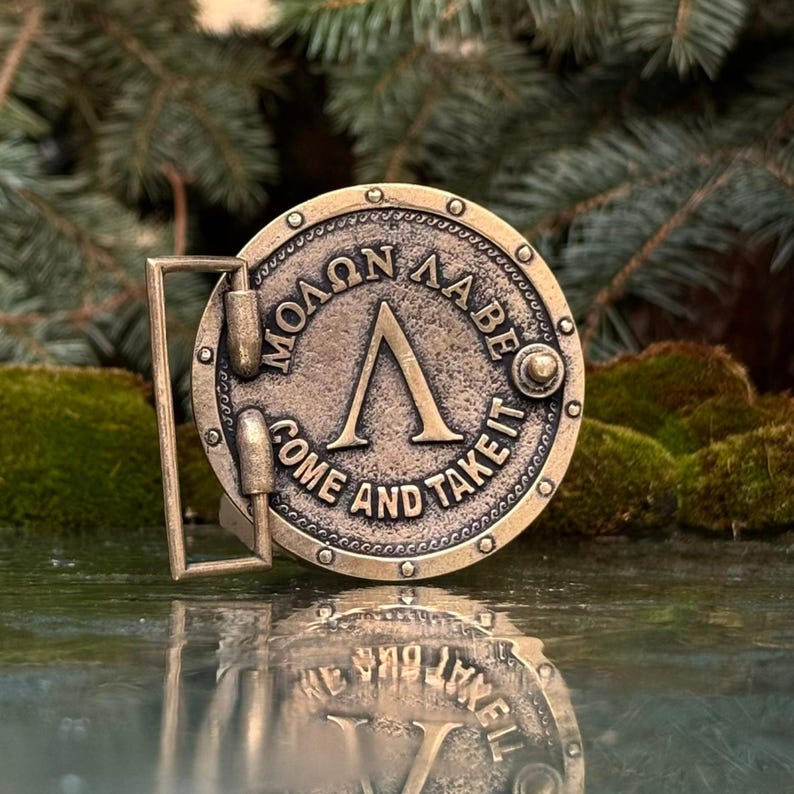 May include: A circular brass-toned metal belt buckle featuring a raised Greek Lambda symbol in the centre. The text "&Mu;&Omicron;&Lambda;&Omega;&Nu; &Lambda;&Alpha;&Beta;&Epsilon;" and "COME AND TAKE IT" is embossed around the perimeter, which is accented with small circular rivets.