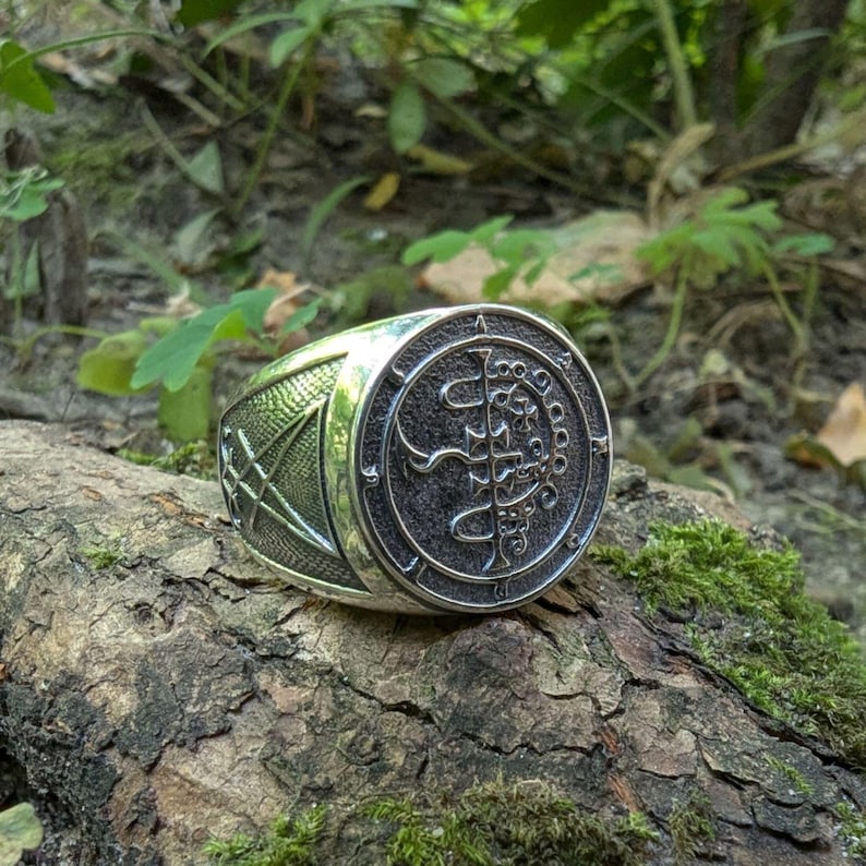 May include: A silver ring with a black sigil design. The sigil is surrounded by a circle of smaller symbols. The ring is set against a background of green foliage.